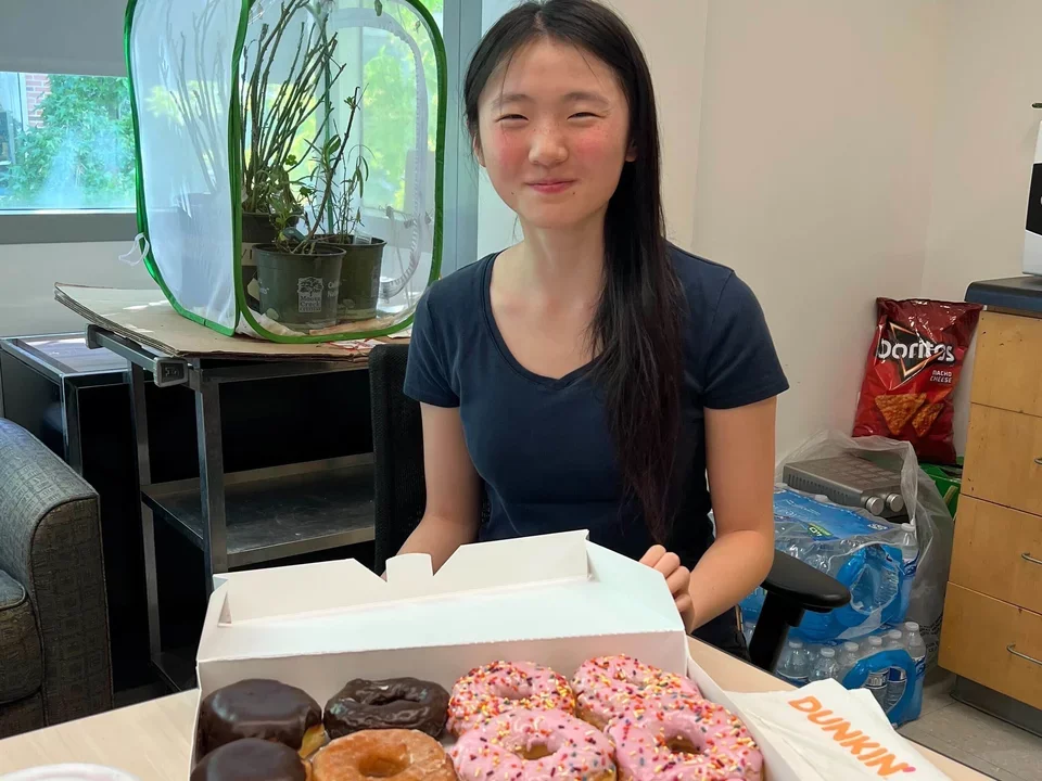 A female lab student is sitting at a table, smiling. A variety box of Dunkin Donuts is open in front of her.