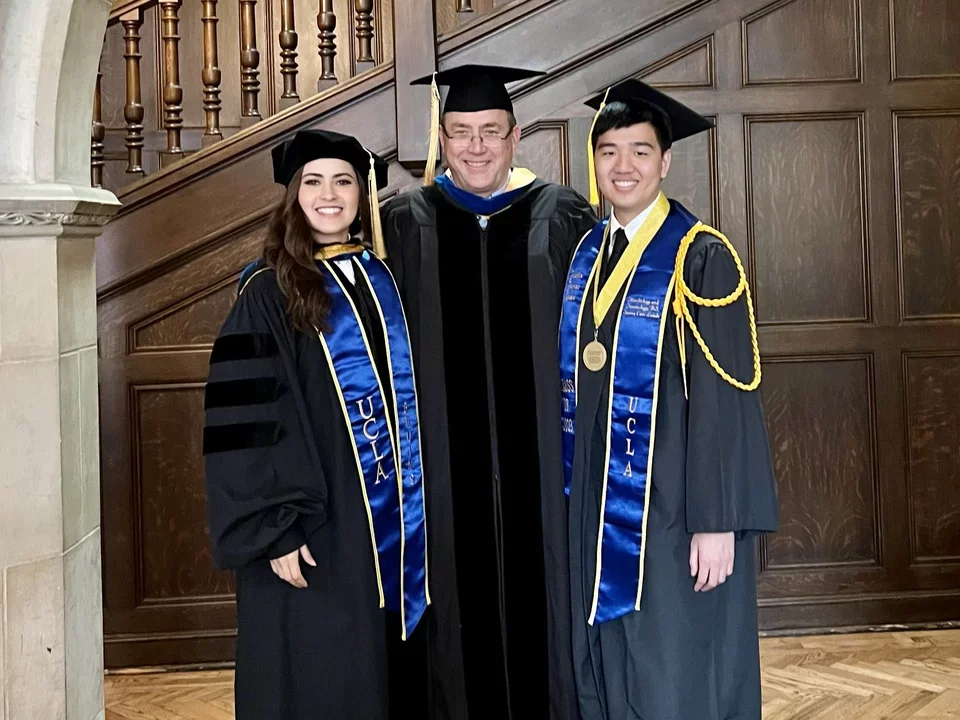 Indoor photo of two students dressed for graduation taking a picture with their PI.