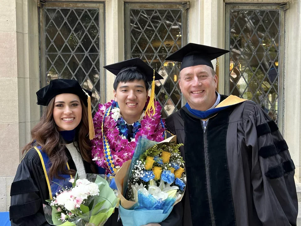 Two students dressed for graduation take a picture with their PI. They are holding flowers in their hands.