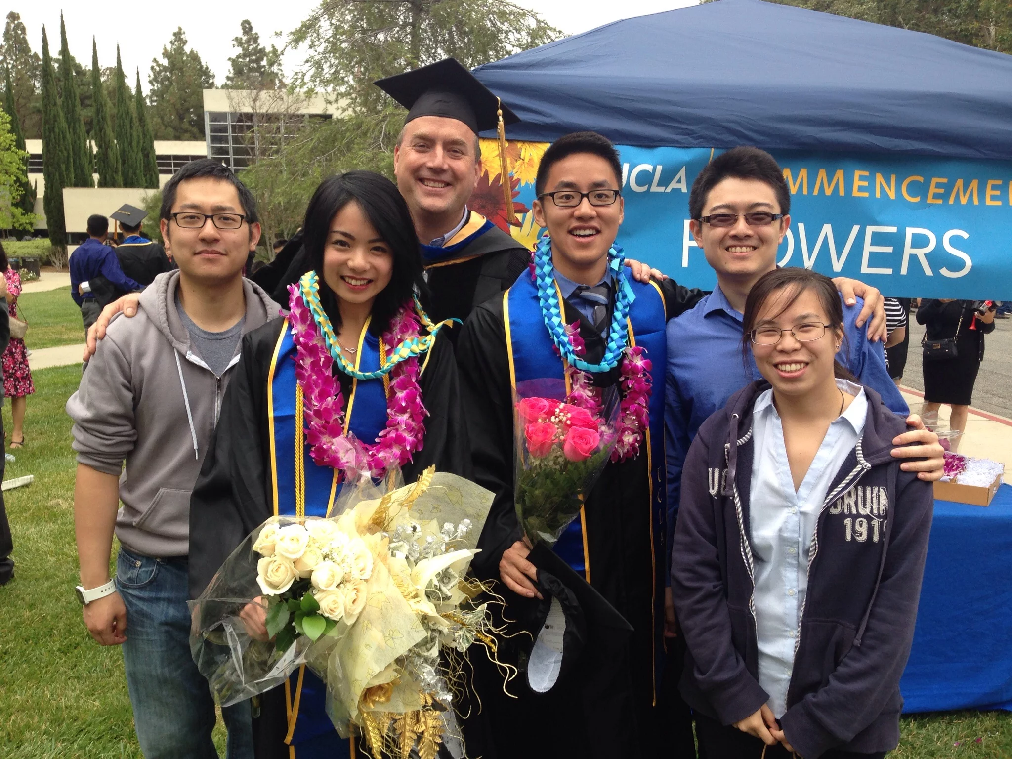 Two lab undergrad students dressed in graduation clothes poses with their family and their PI for a photo.
