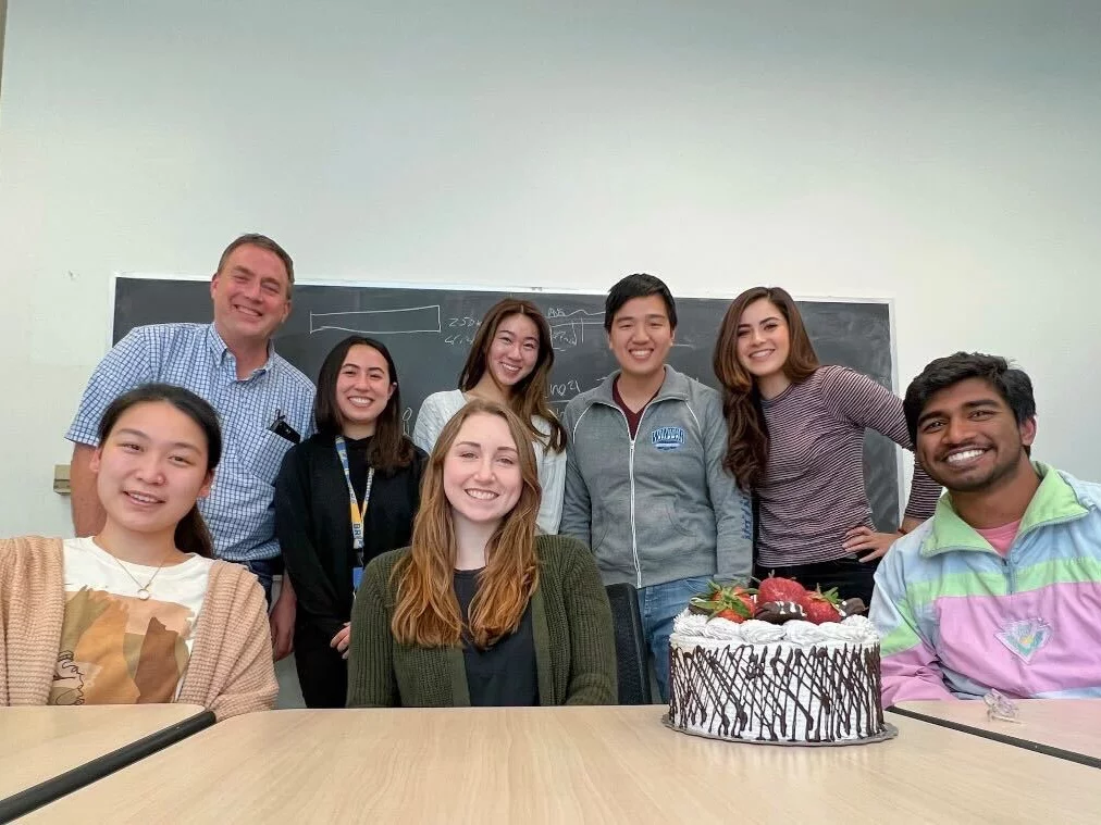 The entire lab celebrates a birthday. They are together in a row with a birthday cake in front of them.