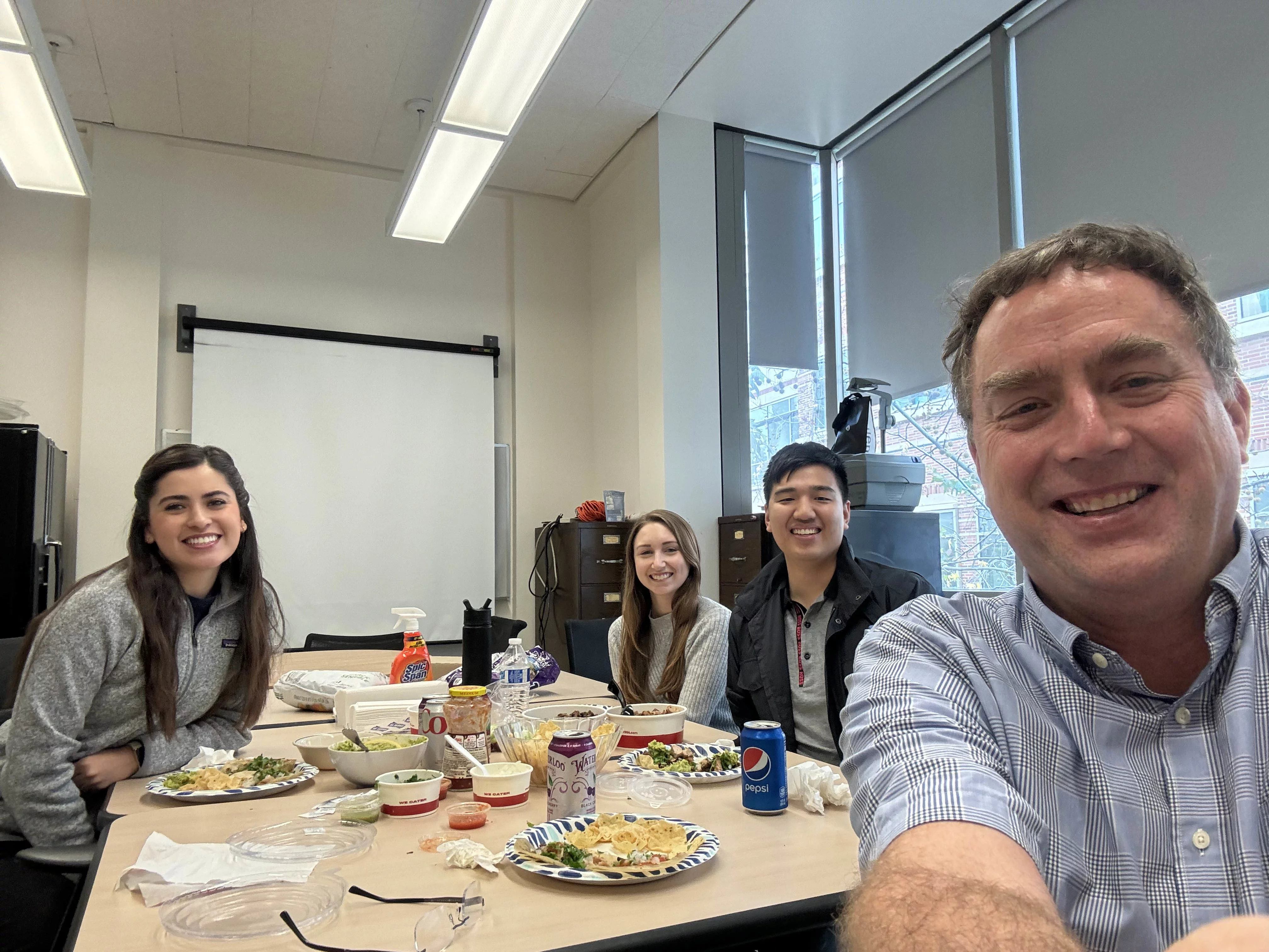 the professor and 3 students taking a selfie in the lab conference room while eating tacos