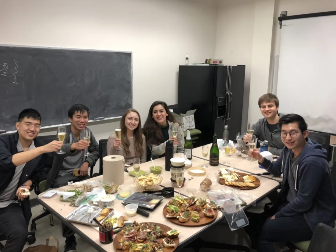 Six lab members gather around the conference room with champagne glasses to celebrate a successful Oral Qualifying Exam.